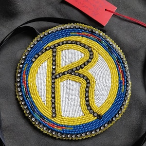 Könnte beinhalten: Ein runder Perlen-Anhänger mit weißem Hintergrund und einem großen gelben Buchstaben "R" in der Mitte. Der Buchstabe ist mit schwarzen Perlen umrandet und von einem blauen und gelben Perlenrand umgeben.