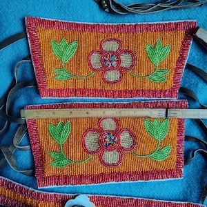 May include: Close-up of beaded Native American crafts. The orange beaded pieces feature a floral design with green leaves and a red border. The central flower has a colorful beaded center. Leather straps and a ruler are also visible.