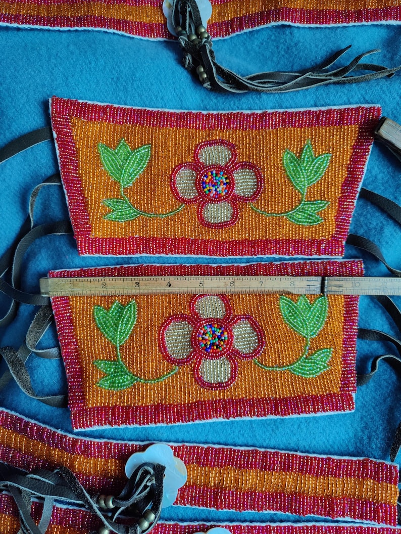 May include: Close-up of beaded Native American crafts. The orange beaded pieces feature a floral design with green leaves and a red border. The central flower has a colorful beaded center. Leather straps and a ruler are also visible.