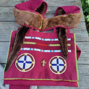 Könnte beinhalten: Eine handgefertigte, bordeauxfarbene Tasche mit Fellakzenten und Perlendetails. Die Tasche hat zwei kreisförmige Designs mit blauen Kreuzen, einer goldenen Fleur-de-Lis und farbigen Perlenstreifen. Die Kanten sind mit grünen und roten Perlen verziert.