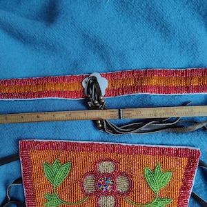 May include: A beaded belt and pouch set with a floral design. The belt is red and orange with a white clasp. The pouch is orange with a red border and a floral design. A wooden ruler is placed next to the belt.
