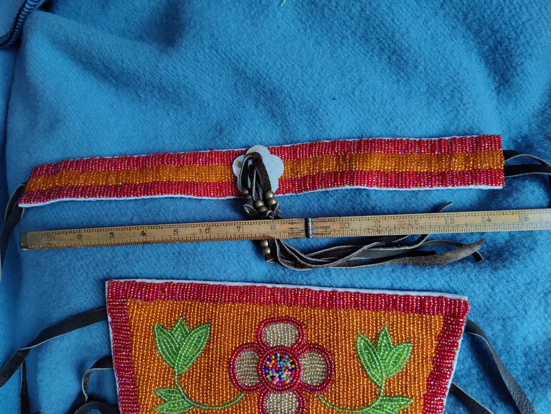 May include: A beaded belt and pouch set with a floral design. The belt is red and orange with a white clasp. The pouch is orange with a red border and a floral design. A wooden ruler is placed next to the belt.