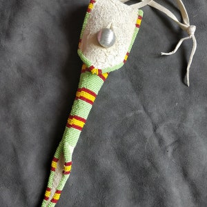 Puede incluir: Una bolsa de cuentas verdes y amarillas con ribete rojo y una solapa de cuero blanco. La bolsa tiene una bola de metal plateado unida a ella y una correa de cuero blanco.