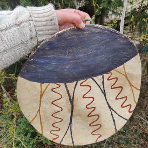 Peut inclure: Une pièce décorative ronde, peinte à la main, avec une section supérieure bleu foncé et une section inférieure beige. La section inférieure présente un motif de zigzags marron et rouges et de lignes courbes noires et jaunes. Une boucle verte est attachée en haut.
