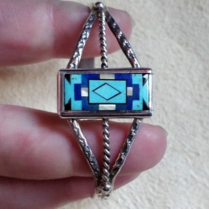 Op de afbeelding: Een zilveren armband met een turquoise en blauw ingelegd ontwerp. De armband is gemaakt van gedraaid zilverdraad en heeft een geometrisch patroon.