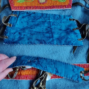 May include: Close-up of a handcrafted textile with intricate beadwork and blue fabric. The design features orange, red, and green beaded floral patterns. Leather straps and decorative elements are also visible, showcasing traditional craftsmanship.