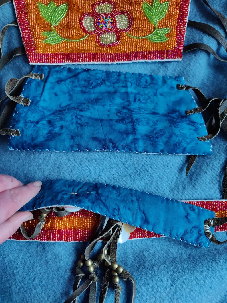May include: Close-up of a handcrafted textile with intricate beadwork and blue fabric. The design features orange, red, and green beaded floral patterns. Leather straps and decorative elements are also visible, showcasing traditional craftsmanship.