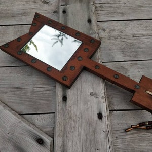 May include: A wooden hand mirror with a rectangular mirror inset and a decorative arrow-shaped handle. The mirror is surrounded by metal studs and the handle has a beaded tassel.