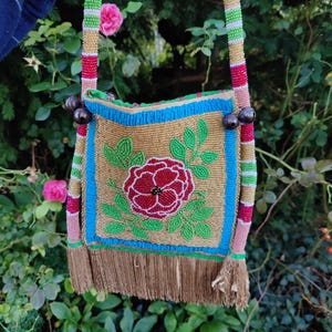 May include: A beaded shoulder bag with a floral design. The bag features a gold background with a red rose and green leaves. The edges are trimmed with blue beads, and the strap is striped with pink, green, and gold beads. The bag has a brown fringe.