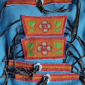 May include: Handmade beaded accessories featuring vibrant orange, red, and green floral designs. The set includes headbands and rectangular pieces, all adorned with intricate beadwork and leather ties. The floral patterns are centered with colorful beads.