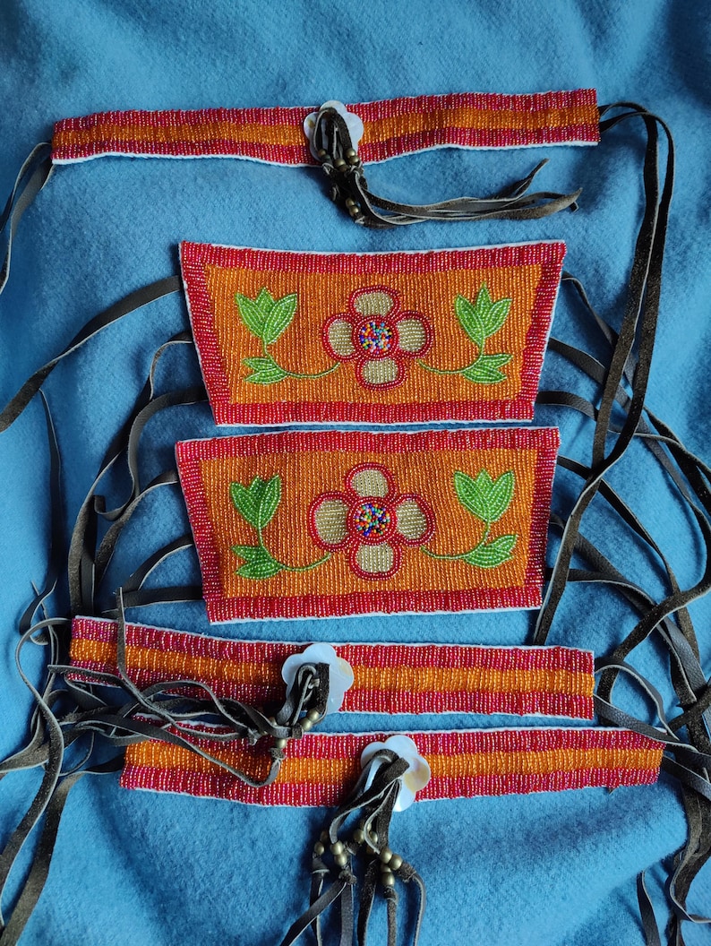 May include: Handmade beaded accessories featuring vibrant orange, red, and green floral designs. The set includes headbands and rectangular pieces, all adorned with intricate beadwork and leather ties. The floral patterns are centered with colorful beads.