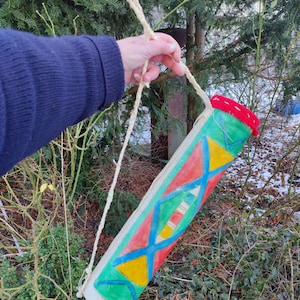 Könnte beinhalten: Eine bunte, zylindrische Stofftasche mit rotem Oberteil und weißem Trageriemen. Die Tasche ist mit geometrischen Formen in Rot-, Gelb-, Blau- und Grüntönen verziert.