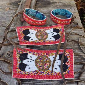 May include: Handmade beaded accessories featuring a red base with intricate designs. The pieces include two bracelets with a blue patterned interior and two rectangular pieces with a central floral design in black, white, and gold. Brown leather straps are attached.