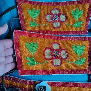May include: Close-up of beaded Native American craftwork. The design features orange and red beaded rectangles with floral patterns in green, red, and gold. The background is a blue fabric.
