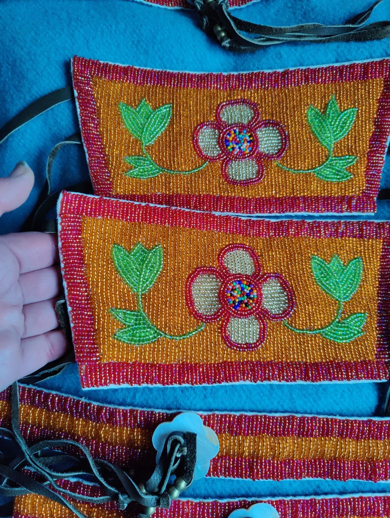 May include: Close-up of beaded Native American craftwork. The design features orange and red beaded rectangles with floral patterns in green, red, and gold. The background is a blue fabric.
