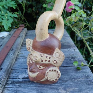 Könnte beinhalten: Eine Keramikflasche in Form eines menschlichen Gesichts mit einem langen Ausguss. Das Gesicht ist in Braun, Weiß und Schwarz mit geometrischen Mustern bemalt. Die Flasche steht auf einer Holzoberfläche.