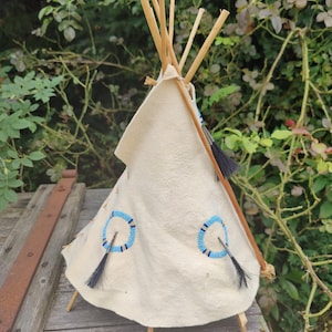 May include: A miniature teepee made of white fabric with blue beaded accents and black feathers. The teepee is supported by wooden poles and is sitting on a wooden surface.