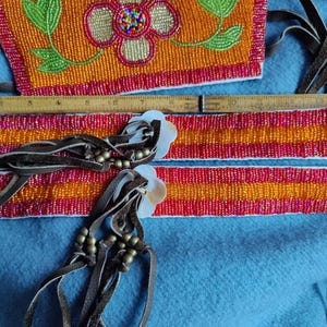 May include: Close-up of Native American beaded crafts. The pieces feature orange, red, and yellow beads with floral and geometric designs. Leather fringe and shell accents add detail. A wooden ruler is also visible.
