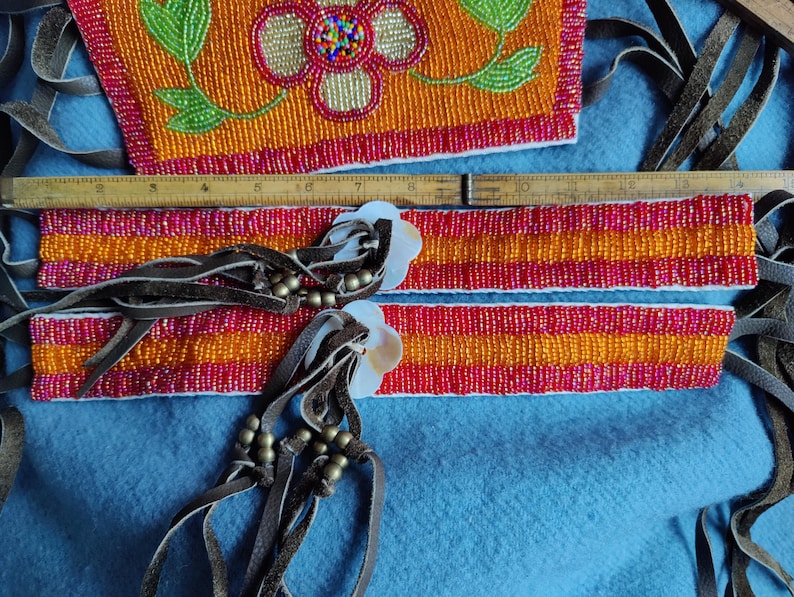 May include: Close-up of Native American beaded crafts. The pieces feature orange, red, and yellow beads with floral and geometric designs. Leather fringe and shell accents add detail. A wooden ruler is also visible.