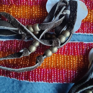 May include: Close-up of a beaded bracelet with red, orange, and gold beads. The bracelet has brown leather straps and gold-colored metal beads. A white, flower-shaped shell is attached to the bracelet.