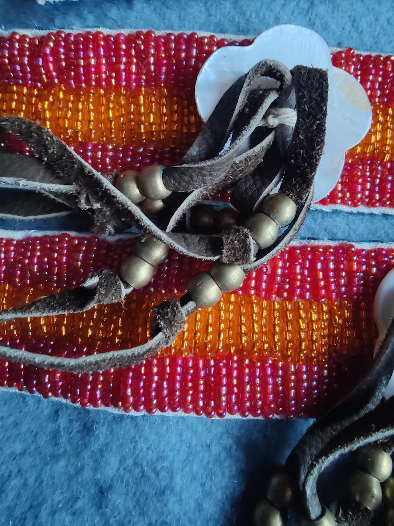 May include: Close-up of a beaded bracelet with red, orange, and gold beads. The bracelet has brown leather straps and gold-colored metal beads. A white, flower-shaped shell is attached to the bracelet.