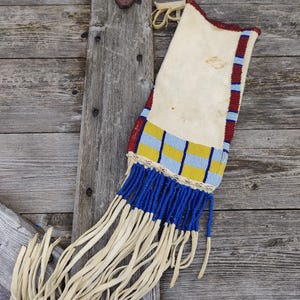 May include: A cream-coloured, fringed pouch with intricate beadwork. The top edge features red, blue, and white beads, while the body showcases blue and yellow geometric patterns. Long, cream-coloured fringe hangs from the bottom.