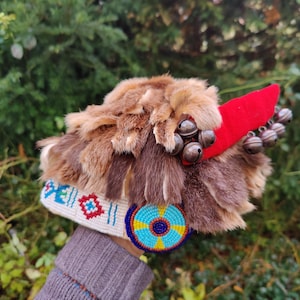 Può includere: Un cappello di pelliccia marrone e bianco con una piuma rossa e campanelli. Il cappello è decorato con perline colorate e un design circolare di perline.