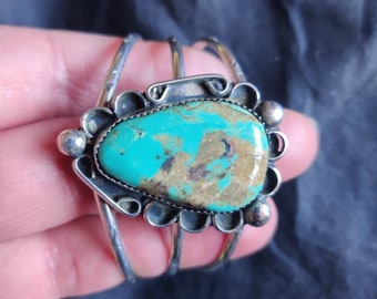 May include: A silver bracelet with a turquoise stone set in a silver bezel. The bracelet has three bands and is decorated with small silver beads.