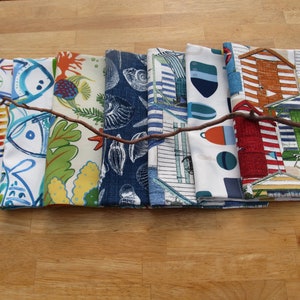 May include: A collection of folded fabric napkins with various coastal-themed patterns. Designs include fish, coral, seashells, beach huts, and nautical elements in shades of blue, green, red, and white. A brown twig lies across the napkins.