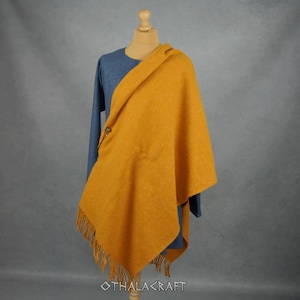 May include: A blue long-sleeved tunic worn under a mustard yellow wool-textured shawl. The shawl features a triangular drape, a fringe along the bottom edge, and is secured at the shoulder with a small metal fastener. The text OTHALACRAFT is visible.