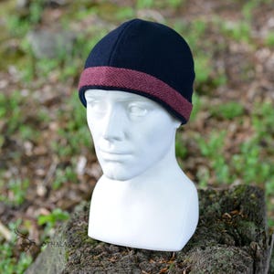 May include: A navy blue beanie with a burgundy patterned band. The hat is displayed on a white mannequin head against a blurred green background. The hat is a stylish accessory.