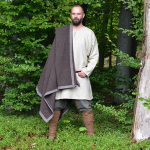 Może przedstawiać: Osoba ubrana w kremową tunikę, szare spodnie i brązowe buty, z brązową szalem z szarym obramowaniem. Osoba stoi w lesie z zielonymi liśćmi.