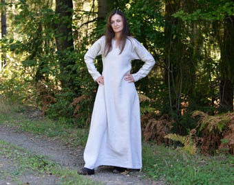 Puede incluir: Un vestido largo hasta los pies con mangas largas en un tejido de color beige claro y tonos naturales. La prenda presenta una silueta sencilla de corte recto con escote redondeado y mangas ajustadas, llegando hasta los tobillos.