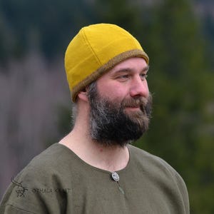 May include: A yellow beanie with a brown trim is worn by a person with a beard. The person is wearing a green shirt. The background is blurred, with trees visible in the distance. The hat appears to be made of wool.