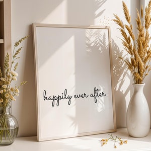 May include: A white framed print with the text "happily ever after" in black cursive script. The print is on a white surface with a vase of dried flowers and a vase of white flowers.