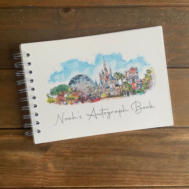 K&ouml;nnte beinhalten: Ein spiralgebundenes Autogrammbuch mit einer Aquarellillustration eines Themenparks. Das Cover zeigt ein Schloss, eine gro&szlig;e Kugel und den Text "Noah's Autograph Book" in Schreibschrift. Auf einer Holzoberfl&auml;che.