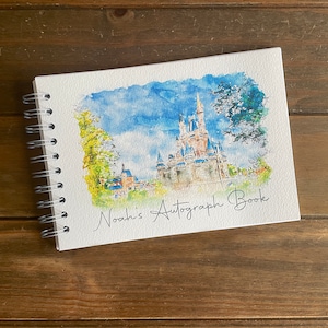 K&ouml;nnte beinhalten: Ein spiralgebundenes Autogrammbuch mit einem Aquarellgem&auml;lde eines Schlosses unter blauem Himmel. Die Worte "Noah's Autograph Book" sind in Kursivschrift geschrieben. Das Buch liegt auf einer Holzoberfl&auml;che.