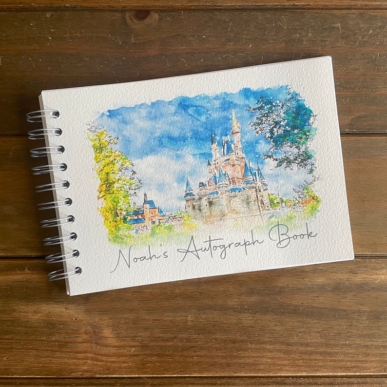 K&ouml;nnte beinhalten: Ein spiralgebundenes Autogrammbuch mit einem Aquarellgem&auml;lde eines Schlosses unter blauem Himmel. Die Worte "Noah's Autograph Book" sind in Kursivschrift geschrieben. Das Buch liegt auf einer Holzoberfl&auml;che.