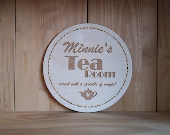 Pode incluir: Uma placa redonda de madeira clara com as palavras "Minnie's Tea Room" recortadas no centro. A placa também inclui a frase "served with a sprinkle of magic!" e um gráfico de bule. A placa tem uma borda decorativa.