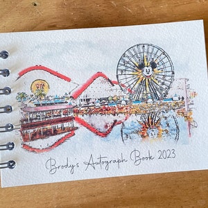 K&ouml;nnte beinhalten: Ein spiralgebundenes Autogrammbuch mit einer Aquarellillustration eines Riesenrads mit Mickey Mouse-Gesicht, Achterbahn und Uferlandschaft. Der Text "Brody's Autograph Book 2023" ist darunter in Kursivschrift geschrieben.