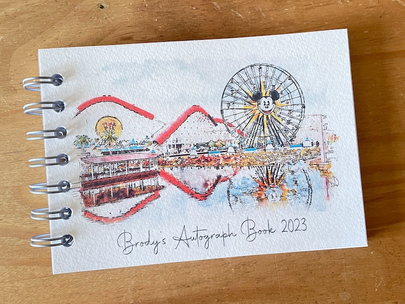 K&ouml;nnte beinhalten: Ein spiralgebundenes Autogrammbuch mit einer Aquarellillustration eines Riesenrads mit Mickey Mouse-Gesicht, Achterbahn und Uferlandschaft. Der Text "Brody's Autograph Book 2023" ist darunter in Kursivschrift geschrieben.