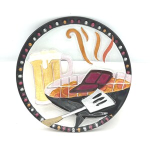 May include: A circular decorative wall plaque featuring a raised relief design of a beer mug with foam, a steak on a grill, and a metal spatula. The border is black with alternating silver and red dots, and orange steam lines rise above the food.