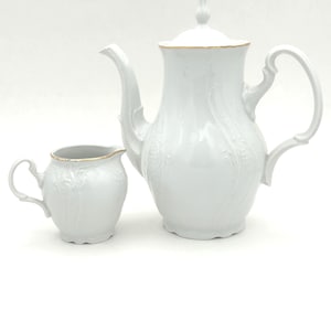 May include: A white porcelain tea set featuring a teapot and creamer. The teapot has a curved spout, a handle, and a lid with a decorative finial. Both pieces have embossed floral patterns and gold trim. The creamer has a small handle and a gold rim.