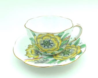 May include: A white porcelain teacup and saucer set featuring a scalloped edge and a gold-trimmed rim. The exterior of the cup and the surface of the saucer are decorated with large yellow flowers, green leaves, and small brown architectural details.