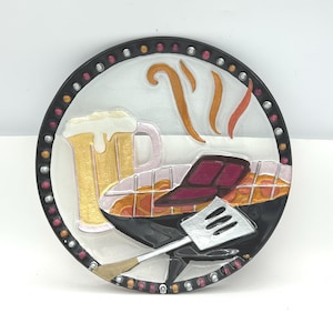 May include: A round decorative plate featuring a barbecue scene. The plate has a black border with small pink and white dots. The image includes a grill, beer mug, spatula, and stylized smoke. The colors are black, white, gold, pink, and orange.