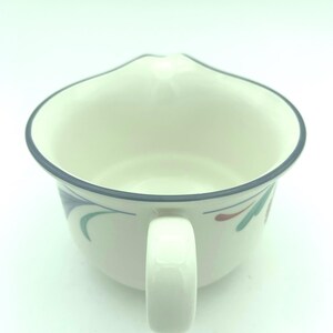Puede incluir: Una jarrita de cer&aacute;mica color crema con un borde azul oscuro y una sola asa. El exterior presenta un patr&oacute;n pintado de formas estilizadas similares a hojas en colores verde azulado y granate. El recipiente tiene un cuerpo ancho y redondeado.