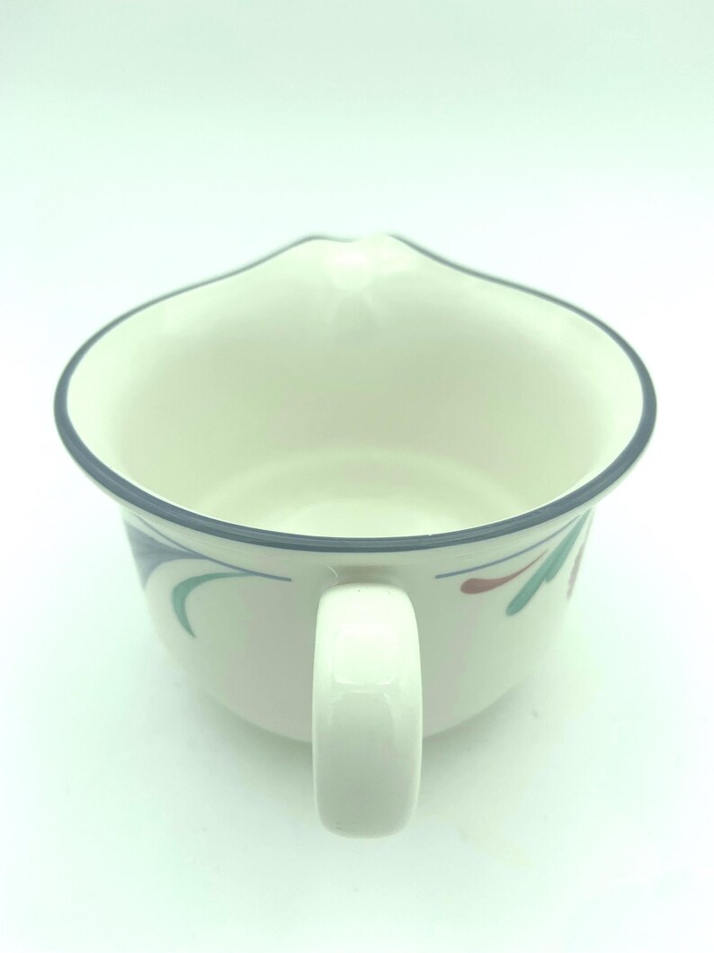 Puede incluir: Una jarrita de cer&aacute;mica color crema con un borde azul oscuro y una sola asa. El exterior presenta un patr&oacute;n pintado de formas estilizadas similares a hojas en colores verde azulado y granate. El recipiente tiene un cuerpo ancho y redondeado.