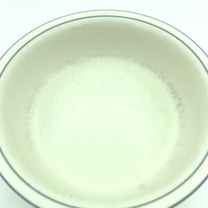 Puede incluir: Un cuenco redondo de cer&aacute;mica de color blanco roto con una sencilla banda fina de color gris oscuro que rodea el borde interior. La superficie interior muestra sutiles signos de desgaste con tenues marcas grises y un borde curvo.
