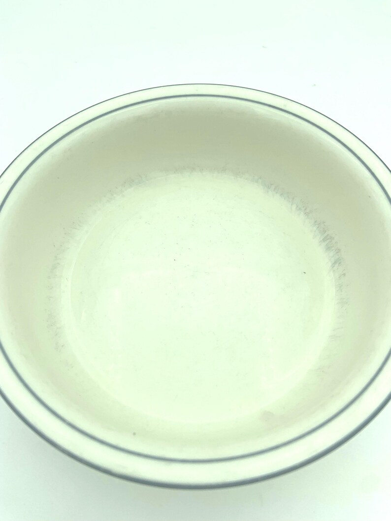 Puede incluir: Un cuenco redondo de cer&aacute;mica de color blanco roto con una sencilla banda fina de color gris oscuro que rodea el borde interior. La superficie interior muestra sutiles signos de desgaste con tenues marcas grises y un borde curvo.