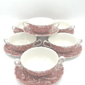 May include: A set of five cream-colored ceramic soup bowls with two handles and matching saucers, each decorated with a detailed red transferware landscape pattern featuring trees, buildings, and pathways around the exterior and rims.